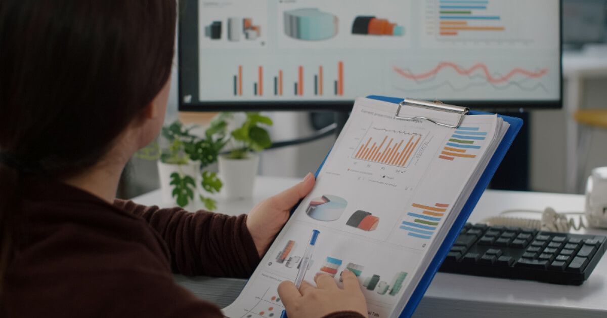 Person holding a clipboard with stats and graphs, overlooking a monitor