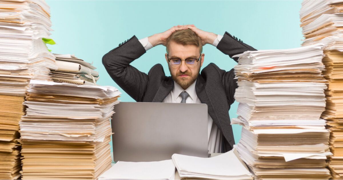 Frustrated man with hands on top of his head behind a laptop and stacks of sheets