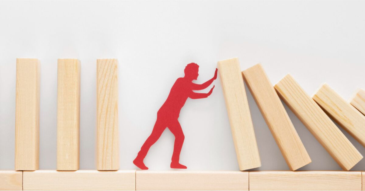 Red paper cutout of a person stopping a series of wooden blocks from falling