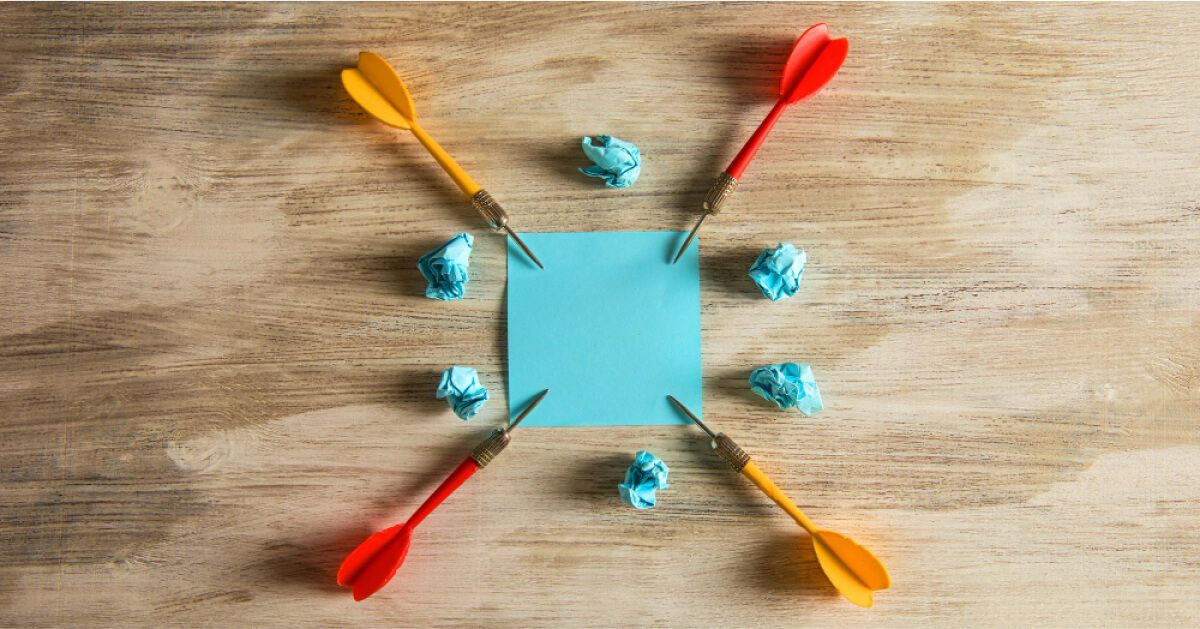 Four darts pointed at a piece of paper, surrounded by balls of crumpled paper