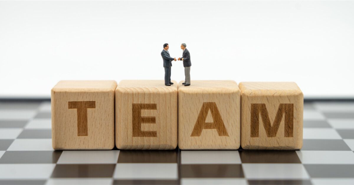 Two miniature businessmen shaking hands over a series of wooden blocks that spell "TEAM"