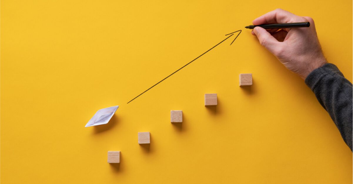 Person drawing an upward moving arrow next to a paper boat and wooden stepped blocks