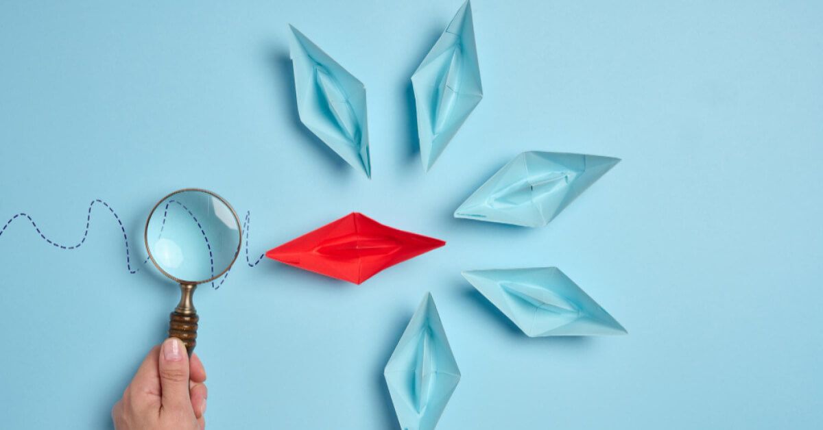 A magnifier looking at a red paper boat with a dotted trail, surrounded by blue paper boats